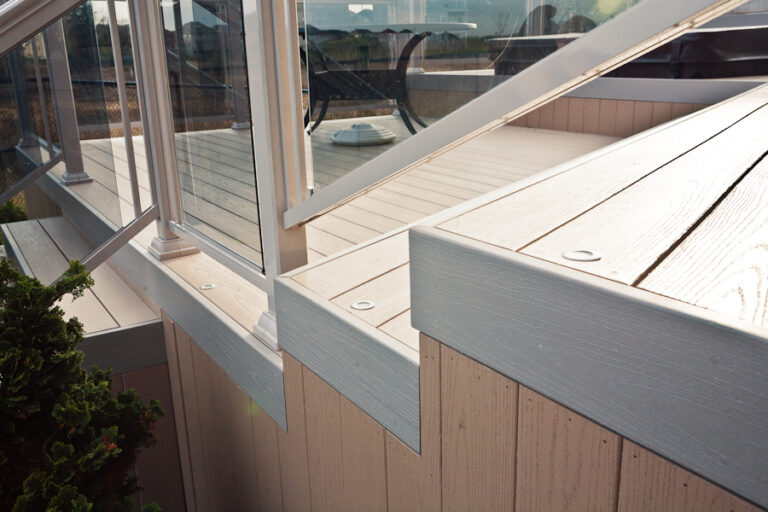 Close-up of a modern deck with light-colored wood, metal railing, and glass panels, seen in daylight.