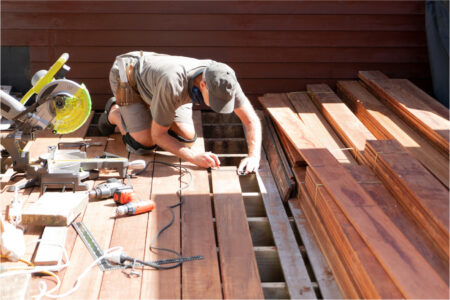 A person wearing a cap and tool belt is kneeling on a wooden deck under construction, measuring with a pencil and tape measure. Various tools, including a miter saw and drill, are nearby, and stacked planks are visible on the right.