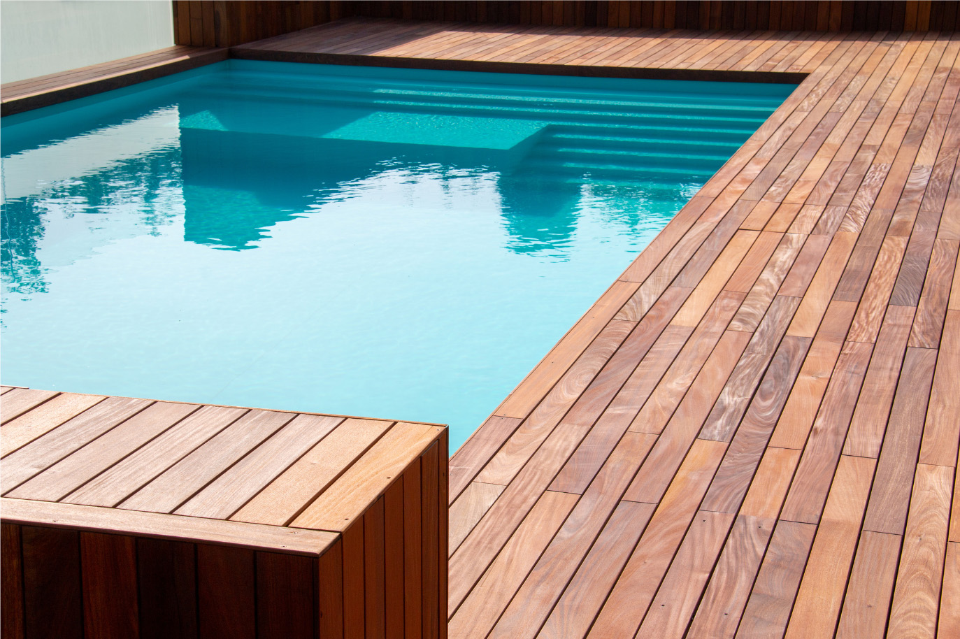 A modern swimming pool with clear blue water, surrounded by a polished wooden deck. Steps lead into the pool, and the deck area is spacious, offering a contemporary and relaxing aesthetic.